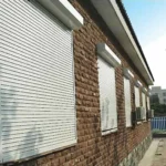A brick building with white rolldown storm shutters closed over its windows; trees and a fence line the paved path beside it. in either North or South Carolina
