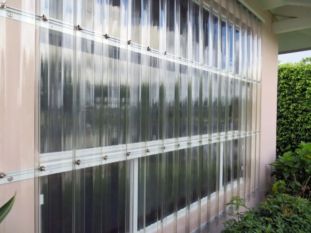 Clear Lexan hurricane panels are installed over a large window on a light-colored building, providing protection while allowing light and visibility; green plants grow nearby. in either North or South Carolina