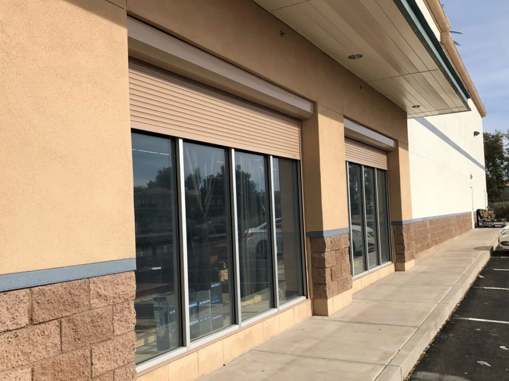 Exterior view of a commercial building with large glass windows, beige and white walls, brown brick accents, and sidewalk beside an empty parking lot—ideal for adding rolldown storm shutters to enhance protection on sunny days. in either North or South Carolina